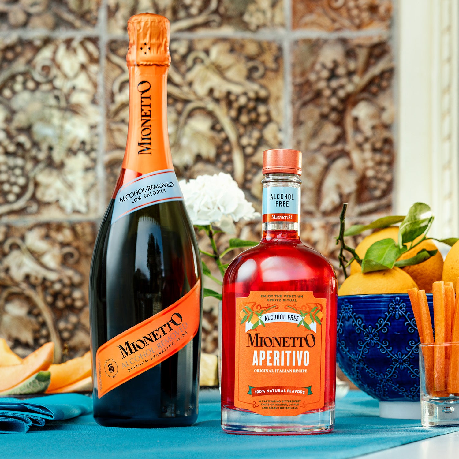 
Two bottles on a blue tablecloth: left, a bottle of Mionetto Alcohol Removed Sparkling Wine; right, a bottle of Mionetto Alcohol Free Aperitivo. In the background, a decorative stone wall, a white flower, and citrus fruits in a bowl are visible.