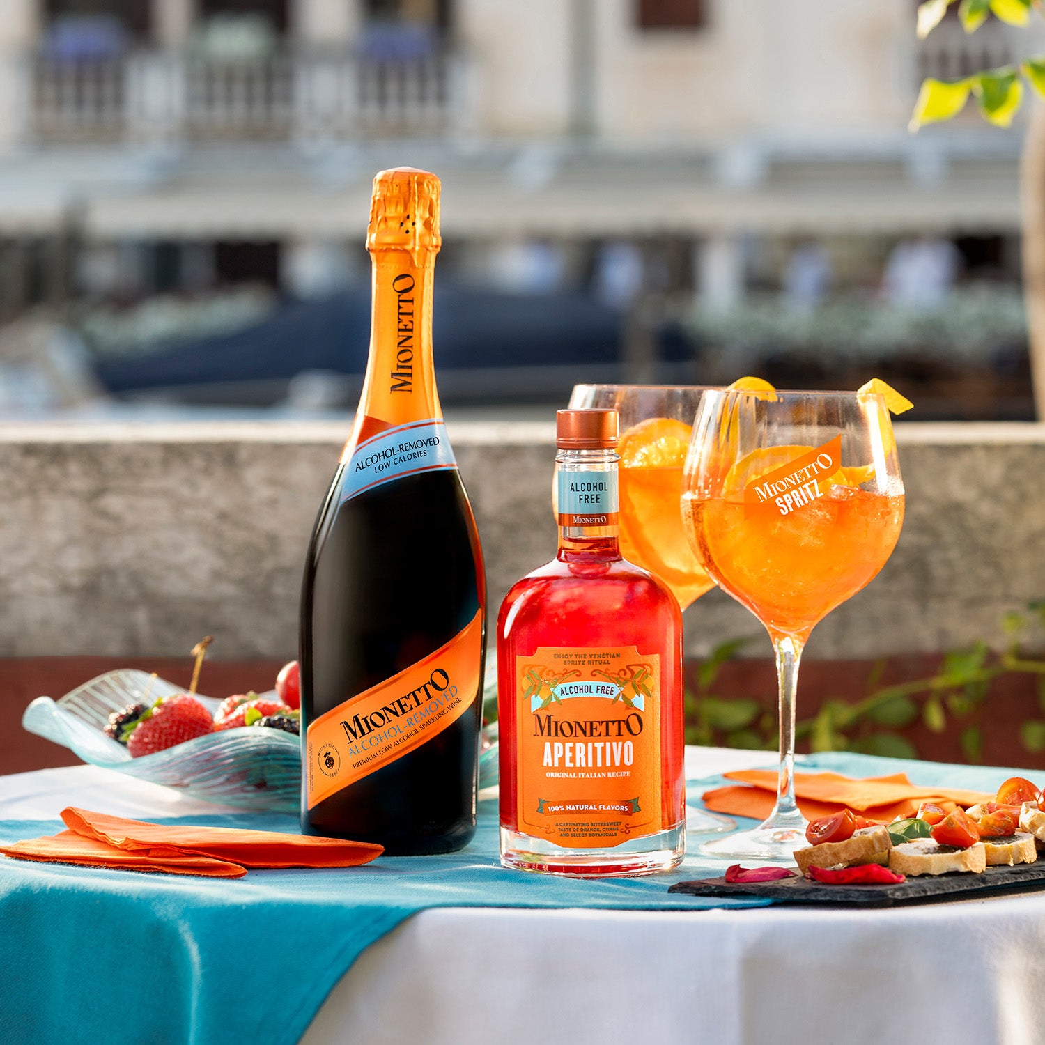 
A table set outdoors with a bottle of Mionetto Alcohol Removed Sparkling Wine, a bottle of Mionetto Alcohol Free Aperitivo, two filled glasses, strawberries, and a platter of appetizers. Theres a turquoise tablecloth and an orange napkin, with a blurred building in the background.