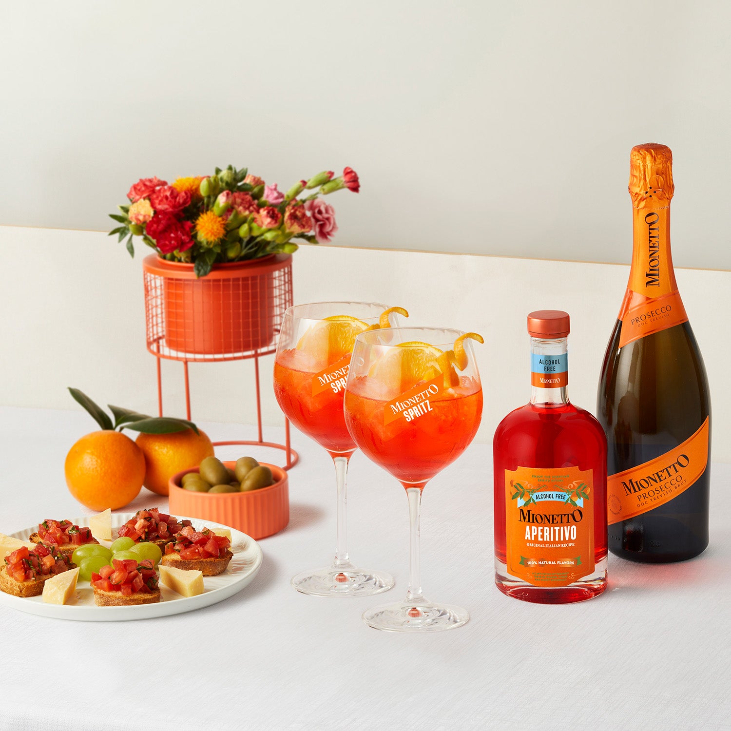 A table setting featuring two glasses of Mionetto Spritzes garnished with orange slices, a bottle of Mionetto  Aperitivo Alcohol Free, a bottle of Mionetto Prosecco, a plate of appetizers, and a vase of colorful flowers.