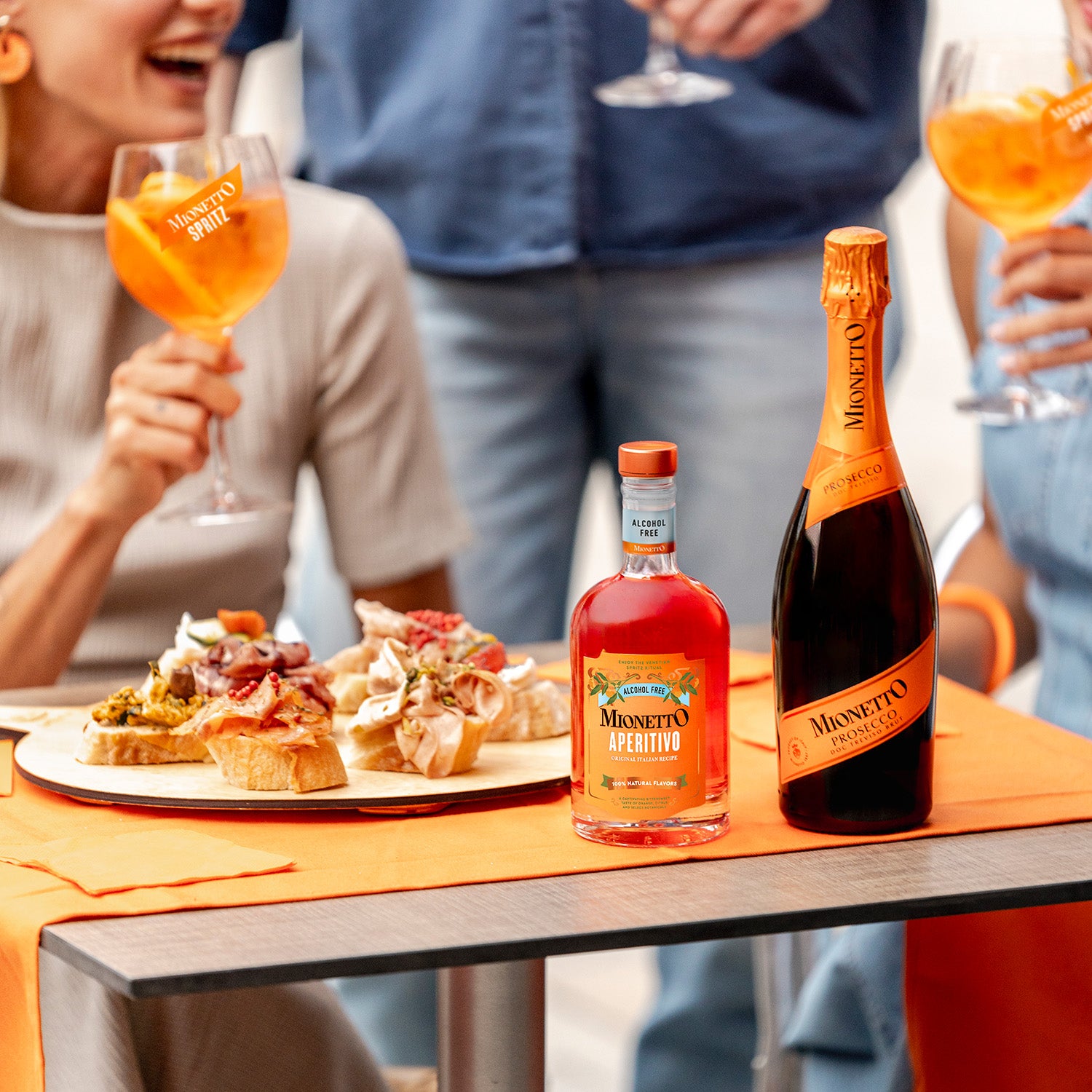 People enjoy Mionetto Spritzes and appetizers around a table. A bottle of Mionetto Aperitivo Alcohol Free and Mionetto Prosecco are on the table. Bright orange tones and an upbeat atmosphere are prominent.